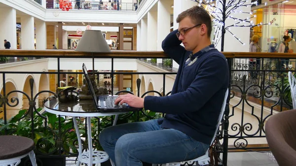 Young man in glasses are sitting at table in shopping mall and working on a laptop — Stock Photo, Image