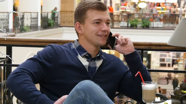 Jonge knappe man koffie drinken en praten over de mobiele telefoon in het café in winkelcentrum. — Stockfoto