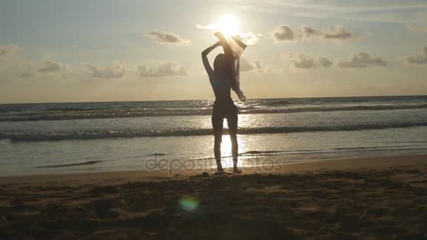 Mladá šťastná žena s šátkem jde pobřeží oceánu při západu slunce. Dívka si léto. Ženská silueta v bikinách s létáním šátek v ruce chůzi na břehu moře. Zpomalený pohyb — Stock video