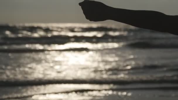 Kvinna hand hälla sea sand genom fingrarna vid solnedgången mot en ocean bakgrund. Arm av ung kvinna med sand beströdde eller falla från den. Grit duggregna från näve av flicka. Närbild — Stockvideo