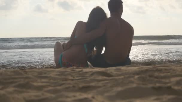 Jovem casal romântico está desfrutando de uma bela vista sentada na praia e abraçando. Uma mulher e um homem sentam-se juntos na areia na praia, admirando o oceano e as paisagens. Fechar — Vídeo de Stock