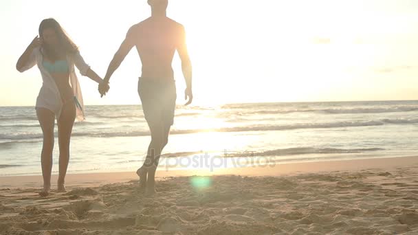 Νεαρό ζευγάρι ευτυχισμένο το περπάτημα στην παραλία και κρατώντας τα χέρια του άλλου στο ηλιοβασίλεμα. Άνδρας και γυναίκα πηγαίνει στην ακτή θάλασσας κατά την όμορφη ανατολή του ηλίου. Αργή κίνηση — Αρχείο Βίντεο