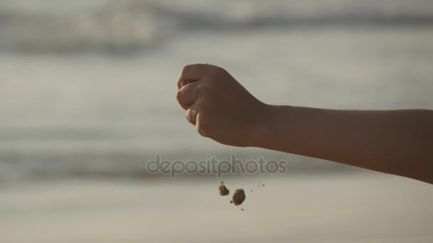 Eine weibliche Hand schüttet bei Sonnenuntergang vor einem Ozeanhintergrund Meeressand durch ihre Finger. Arm einer jungen Frau mit Sand bestreut oder heruntergefallen. Es nieselt aus Mädchenfaust. Nahaufnahme — Stockvideo