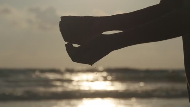 Siluett av kvinna hand hälla sea sand genom hennes fingrar vid solnedgången mot en ocean bakgrund. Arm av ung kvinna med sand beströdde eller falla från den. Grit duggregna från näve av flicka. Närbild — Stockvideo