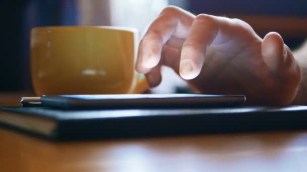 Primer plano de las manos del hombre utilizando el teléfono inteligente para la mensajería. El teléfono descansa en un cuaderno y está al lado de una taza de café — Vídeo de stock