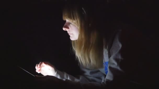 Young handsome blonde girl using her tablet laying on the bed in her lights off house — Stock Video