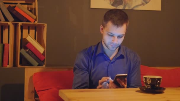 若いハンサムな男は一杯のコーヒーのためのカフェに座っているし、スマート フォンを使用して — ストック動画