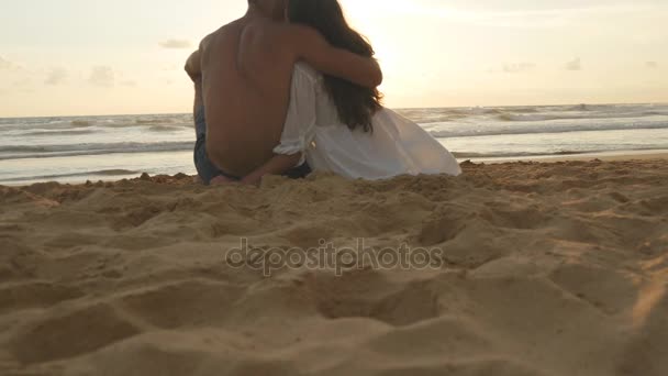 Jeune couple romantique profite d'un magnifique coucher de soleil assis sur la plage et câlins. Une femme et un homme sont assis ensemble dans le sable au bord de la mer, admirant l'océan et les paysages. Gros plan — Video