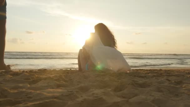 Egy nő és egy férfi ül össze a homok, a tenger partján, megcsodálta az óceán és a táj. Fiatal szerelmespár élvezi gyönyörű naplemente ül a strandon, és átölelte. Közelről — Stock videók