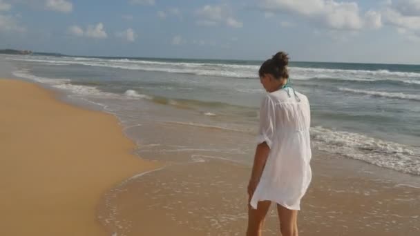 Hermosa mujer en traje de baño y camisa caminando en la playa del mar y las manos levantadas. Chica joven que va a la orilla del mar y disfrutar del verano. Ondas marinas al fondo. Concepto de vacaciones Vista trasera trasera — Vídeo de stock