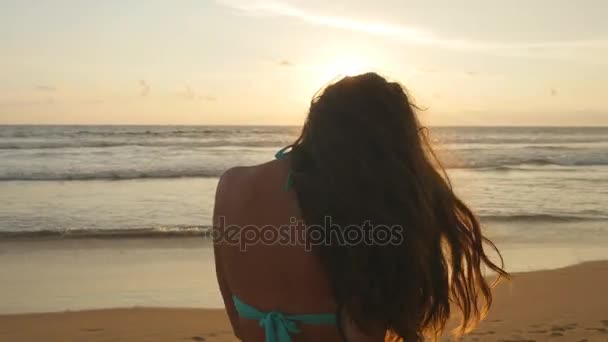 Hermosa joven en bikini de pie cerca del mar al atardecer. Atractiva chica sexy con el pelo largo posando en la orilla del océano al amanecer. Mujer en la playa disfrutando de la vida durante las vacaciones. De cerca. — Vídeo de stock
