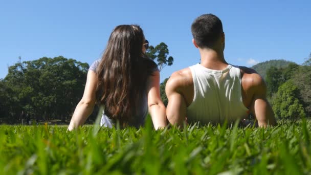 Mladý pár, ležící na zelené trávě v parku a relaxaci. Muž a žena sedí na louce v přírodě a líbání. Dívka a chlapec při pohledu na krajinu a užívat si dovolenou. Zadní zpět nízký úhel pohledu — Stock video