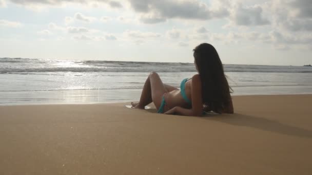 Mulher bonita em biquíni deitado na areia dourada na praia do mar e relaxante durante as férias de verão. Menina bronzeada tomando banho de sol em perfeita costa paradisíaca. Ondas oceânicas ao fundo. Fechar — Vídeo de Stock