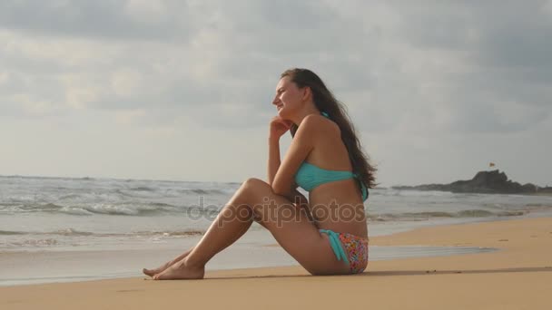 海のビーチの黄金の砂の上に座ってビキニの美しい若い女性。夏の休暇旅行の間に完璧な楽園の海岸でリラックスできる日焼けした少女。背景に海の波。クローズ アップ — ストック動画