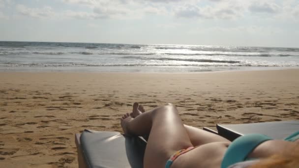 Mujer joven tumbada en el solarium junto al mar y bronceándose. Cuerpo femenino en chaise-longue relajante y disfrutando durante las vacaciones de verano en la playa vacía de arena del océano. Chica en bikini en un resort. De cerca. — Vídeo de stock