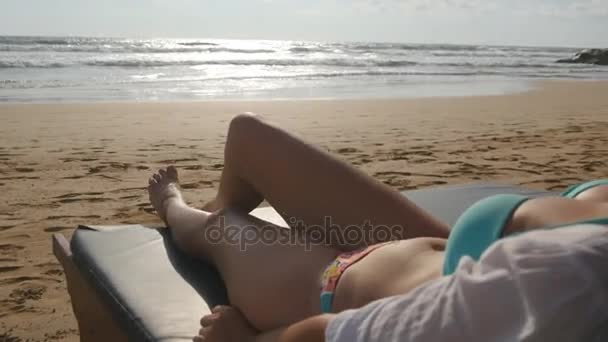 Jonge vrouw liggend op zonnebank door de zee en zonnen. Vrouwelijk lichaam op chaise-longue ontspannen en genieten tijdens de zomervakantie op lege zand oceaan strand. Meisje in bikini op een resort. Close-up — Stockvideo