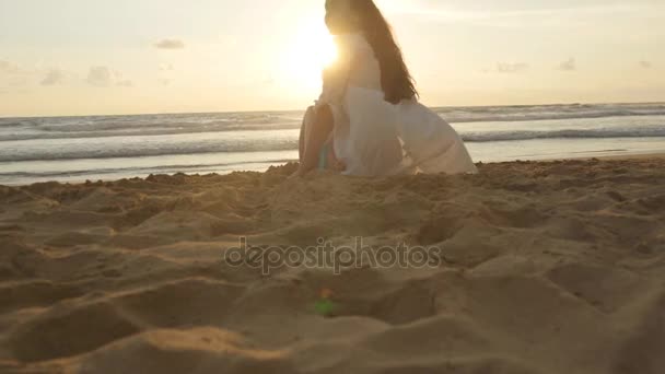 Όμορφη νεαρή γυναίκα που κάθεται στη χρυσή άμμο στην παραλία της θάλασσας κατά τη διάρκεια του ηλιοβασιλέματος και οι κλήσεις προς τον εαυτό του. Κορίτσι χαλάρωση στην ακτή ωκεανό κατά τη διάρκεια των θερινών διακοπών ταξιδεύουν στο ηλιοβασίλεμα. Κύματα του ωκεανού στο παρασκήνιο. Κοντινό πλάνο — Αρχείο Βίντεο