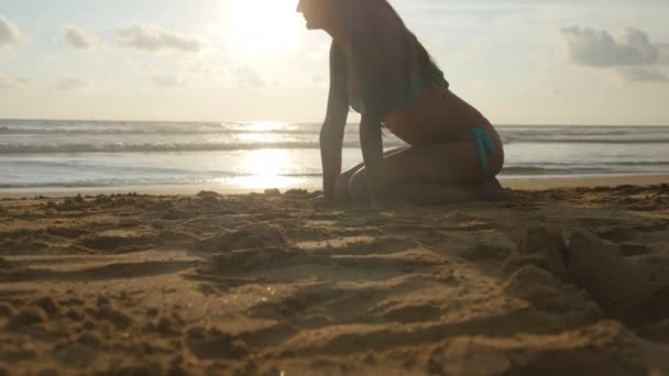 Gyönyörű fiatal nő a bikini, arany homokos tengerparttól a naplemente alatt ül. Lány pihentető nyaralás alatt tökéletes paradicsomi partján utazási napkeltekor. Óceán hullámai háttér. Közelről — Stock videók