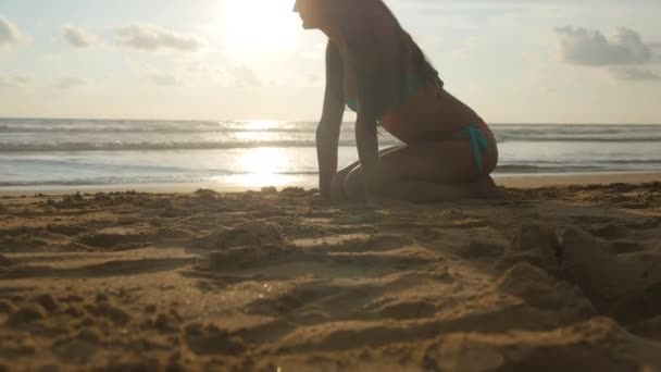 Wanita muda cantik dengan bikini duduk di pasir emas di pantai laut saat matahari terbenam. Gadis santai di pantai surga yang sempurna selama liburan musim panas saat matahari terbit. Gelombang laut di latar belakang. Tutup. — Stok Video