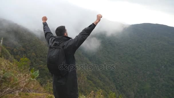 Joven excursionista masculino con mochila que alcanza la cima de la montaña y levanta las manos. Turista hombre de pie en el borde del hermoso cañón, victoriosamente extendiendo los brazos hacia arriba. Vista trasera trasera en cámara lenta — Vídeos de Stock