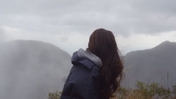 Frau Reisenden sitzt am Rande der schönen Schlucht und Blick auf die Natur. junge Touristinnen im Regenmantel genießen die Aussicht in die Berge. Seitenansicht aus nächster Nähe — Stockvideo