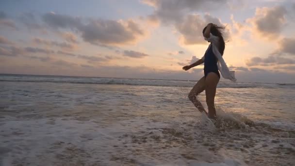 Mujer feliz en traje de baño y camisa caminando en la playa al atardecer. Las olas del océano salpicando los pies femeninos. Joven hermosa chica disfrutando de la vida y divertirse en la orilla del mar. Vacaciones de verano. Movimiento lento — Vídeo de stock