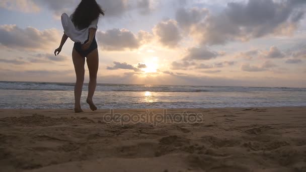 Young happy woman in swimsuit and shirt going to the water at the ocean beach at sunset. Girl enjoying summer vacation or holiday. Female in bikini walking and jogging on the sea shore. Slow motion — Stock Video