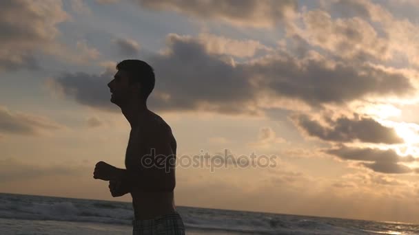 Young sporty man running on the sea beach at sunset. Athletic guy jogging along ocean shore during sunrise. Male sportsman exercising outdoor. Healthy active lifestyle. Workout at nature. Slow motion — Stock Video