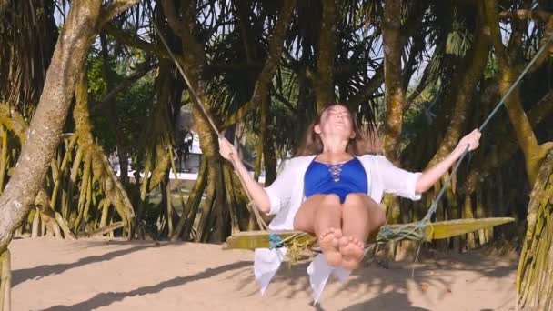 Young happy woman in swimsuit and shirt relaxing at swing at tropical ocean beach. Beautiful girl sitting on swing and enjoying summer vacation or holiday. Female having fun at sea shore. Slow motion — Stock Video