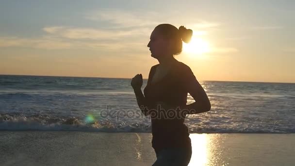 Silhouet van jonge vrouw draait op zee strand bij zonsondergang. Meisje joggen langs Oceaan kust tijdens zonsopgang. Vrouwelijke sportman uit te oefenen buiten. Gezonde actieve training levensstijl op aard. Slow motion — Stockvideo