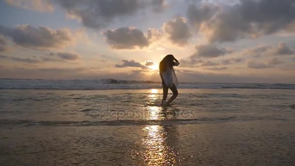 Ευτυχισμένη γυναίκα με μπικίνι και πουκάμισο περπάτημα στο νερό στον ωκεανό στην παραλία το ηλιοβασίλεμα. Όμορφη κοπέλα απολαμβάνοντας διακοπές και διασκέδαση στη θάλασσα ακτή στην Ανατολή του ηλίου. Καλοκαιρινές διακοπές. Αργή κίνηση — Αρχείο Βίντεο