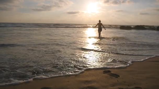 Νεαρή γυναίκα με μαγιό πηγαίνει στην παραλία της θάλασσας, να κολυμπά στο ηλιοβασίλεμα. Όμορφη νεαρή κοπέλα περπάτημα στην ακτή προς τον ωκεανό κατά τη διάρκεια διακοπών. Χαλαρώστε στις διακοπές το καλοκαίρι. Πίσω πίσω όψη αργή κίνηση — Αρχείο Βίντεο