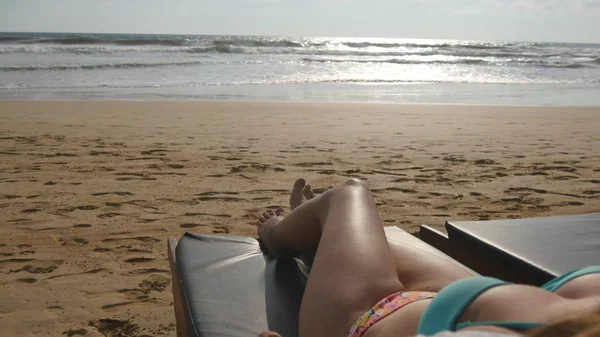 Corpo femminile su chaise-longue rilassante e godendo durante le vacanze estive sulla spiaggia sabbiosa vuota dell'oceano. Giovane donna sdraiata sul lettino in riva al mare e abbronzata. Ragazza in bikini in un resort. Da vicino. — Foto Stock