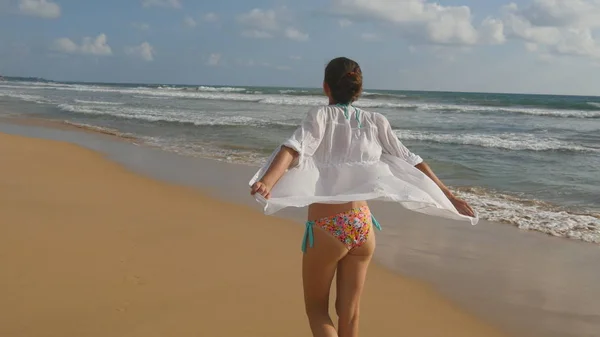 Hermosa mujer en traje de baño y camisa caminando en la playa del mar y las manos levantadas. Chica joven que va a la orilla del mar y disfrutar del verano. Ondas marinas al fondo. Concepto de vacaciones Vista trasera trasera — Foto de Stock