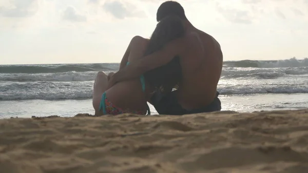 Giovane coppia romantica sta godendo bella vista seduta sulla spiaggia e abbracci. Una donna e un uomo si siedono insieme nella sabbia sulla riva del mare, ammirando l'oceano e i paesaggi. Primo piano — Foto Stock
