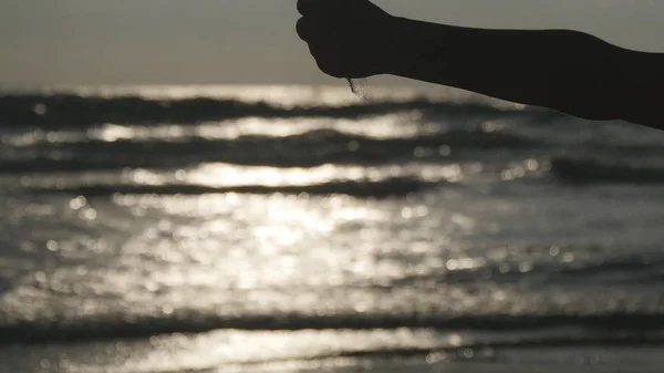 Eine weibliche Hand schüttet bei Sonnenuntergang vor einem Ozeanhintergrund Meeressand durch ihre Finger. Arm einer jungen Frau mit Sand bestreut oder heruntergefallen. Es nieselt aus Mädchenfaust. Nahaufnahme — Stockfoto