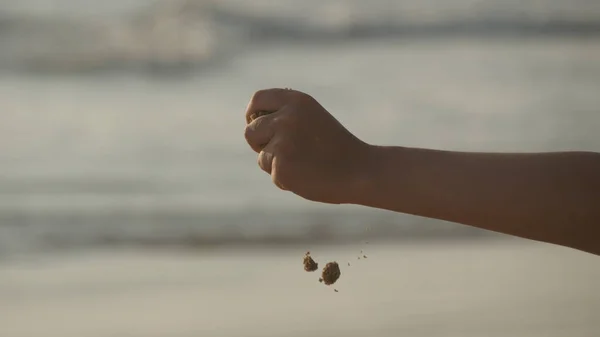 Eine weibliche Hand schüttet bei Sonnenuntergang vor einem Ozeanhintergrund Meeressand durch ihre Finger. Arm einer jungen Frau mit Sand bestreut oder heruntergefallen. Es nieselt aus Mädchenfaust. Nahaufnahme — Stockfoto