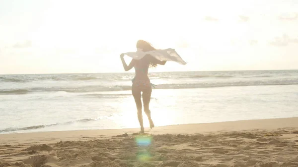 Gelukkig jongedame met sjaal bij zonsondergang op het strand van de oceaan uitgevoerd. Meisje geniet zomer. Vrouw in bikini met vliegende sjaal in handen joggen op de kust. Close-up — Stockfoto