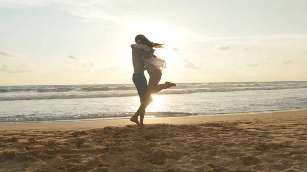 Jeune couple court sur la plage, homme câlin et tourner autour de sa femme au coucher du soleil. La fille saute dans les bras de son petit ami, il la tourbillonne au bord de la mer magnifique. S'amuser ensemble en vacances. Gros plan — Photo