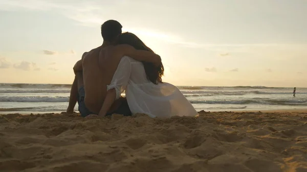 Una donna e un uomo si siedono insieme nella sabbia sulla riva del mare, ammirando l'oceano e i paesaggi. Giovane coppia romantica sta godendo bel tramonto seduto sulla spiaggia e abbracci. Da vicino. — Foto Stock