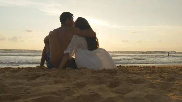 Giovane coppia romantica sta godendo bel tramonto seduto sulla spiaggia e abbracci. Una donna e un uomo si siedono insieme nella sabbia sulla riva del mare, ammirando l'oceano e i paesaggi. Da vicino. — Foto Stock