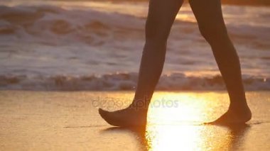 Kadın ayakları çıplak ayakla günbatımında deniz kıyısında yürüyüş. Genç kadın okyanus plaj boyunca gündoğumu sırasında bacaklar. Shoreline, sığ suda adım kız. Yaz tatili. Ağır çekim yakın çekim