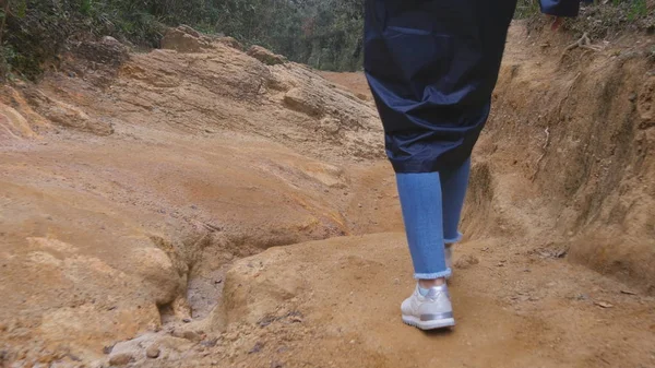 Siguiendo a la turista femenina en impermeable caminando por el sendero de piedra. Pies de mujer caminando a través de un estrecho cañón. Piernas de chica en zapatillas de deporte que van en el camino rocoso. Bajo ángulo de visión Primer plano — Foto de Stock