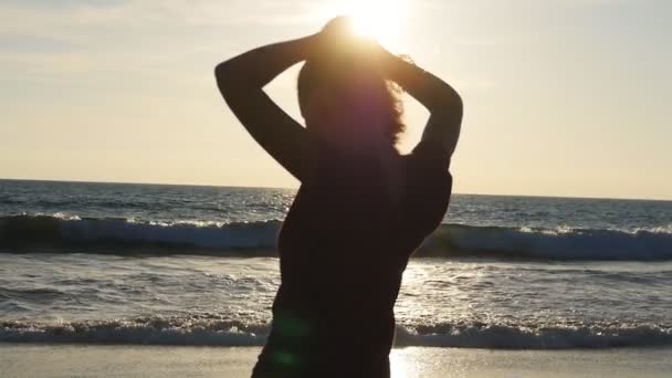 Mujer morena joven atando cola de caballo en la playa cerca del mar al atardecer. Hermosa chica exprimir su cabello en la orilla del océano al amanecer. Mujer turista trenzando el cabello y haciendo peinado. Cuidado del cabello cámara lenta — Vídeos de Stock