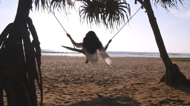 Jovem mulher feliz em maiô e camisa relaxante no balanço na praia do oceano tropical. Menina bonita sentado no balanço e desfrutar de férias de verão ou férias. A fêmea diverte-se na costa. Visão traseira traseira — Vídeo de Stock