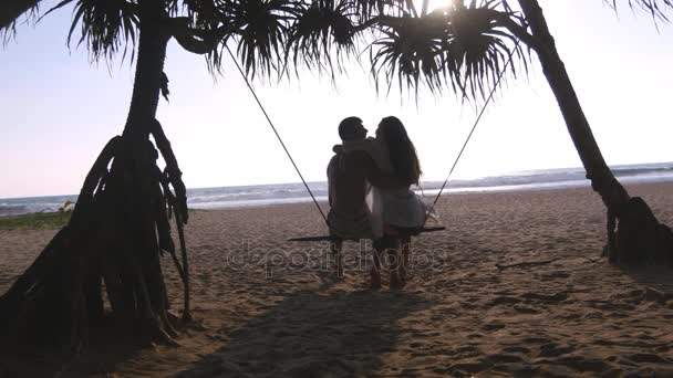 Jovem casal romântico sentados juntos no balanço na praia do mar tropical e abraçando. Par no amor relaxante e beijando no oceano costa exótica durante as férias de verão. Conceito de férias Visão traseira traseira — Vídeo de Stock