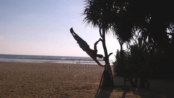 Young guy demonstrates human flag at sea beach. Athletic man doing gymnastics elements on palm tree at exotic ocean shore. Male sportsman performs strength exercises during workout. Training outside — Stock Video