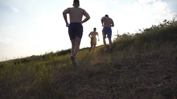 Fiatal férfiak csoportja fut fel a zöld dombon a kék ég felett napkitöréssel a háttérben. A férfi sportolók kocognak a természetben naplementekor. A sportfutók napkeltekor felmennek a dombra. Lassú mozgás. — Stock videók