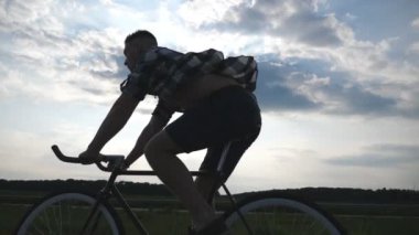 Genç adam arka plan güzel günbatımı gökyüzüne vintage bisiklet sürme silüeti. Sportif adam ülkede yol Bisiklete binme. Erkek bisikletçi sabit binicilik bisiklet dişli. Sağlıklı aktif yaşam tarzı Slowmo