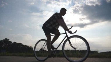 Genç adam arka plan güzel günbatımı gökyüzüne vintage bisiklet sürme silüeti. Adam ülkede yol Bisiklete binme. Erkek bisikletçi sabit binicilik bisiklet dişli. Sağlıklı aktif yaşam tarzı ağır çekim